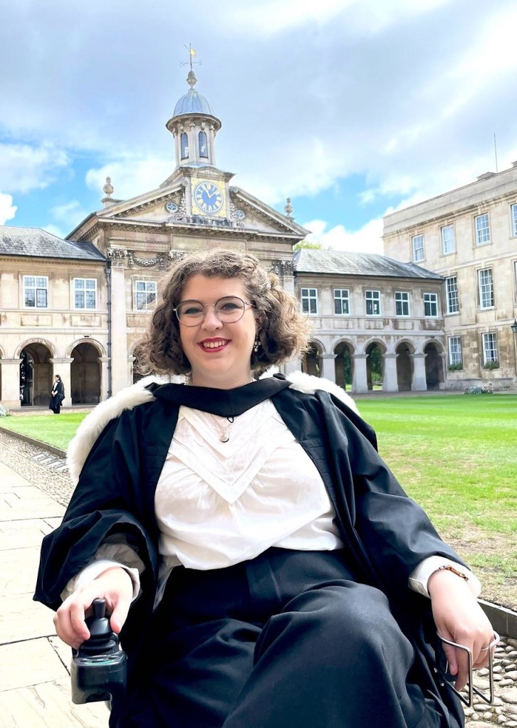 A photograph of Anna, a white woman with short brown hair. Anna is wearing graduate robes and is pictured outside a university college building in an electric wheelchair, smiling. 