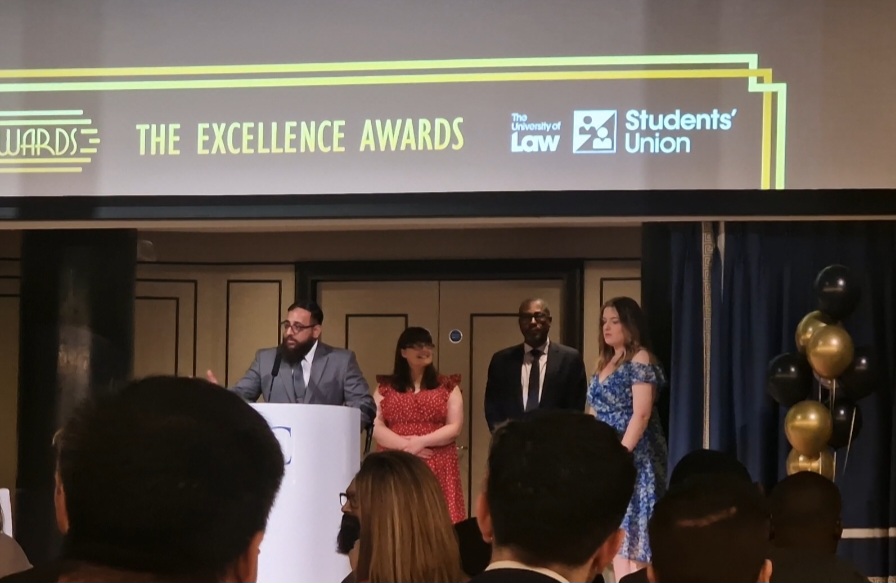 Wasim stood at a speaker podium wearing a suit, with three people behind and a sign above the head which reads the excellence awards