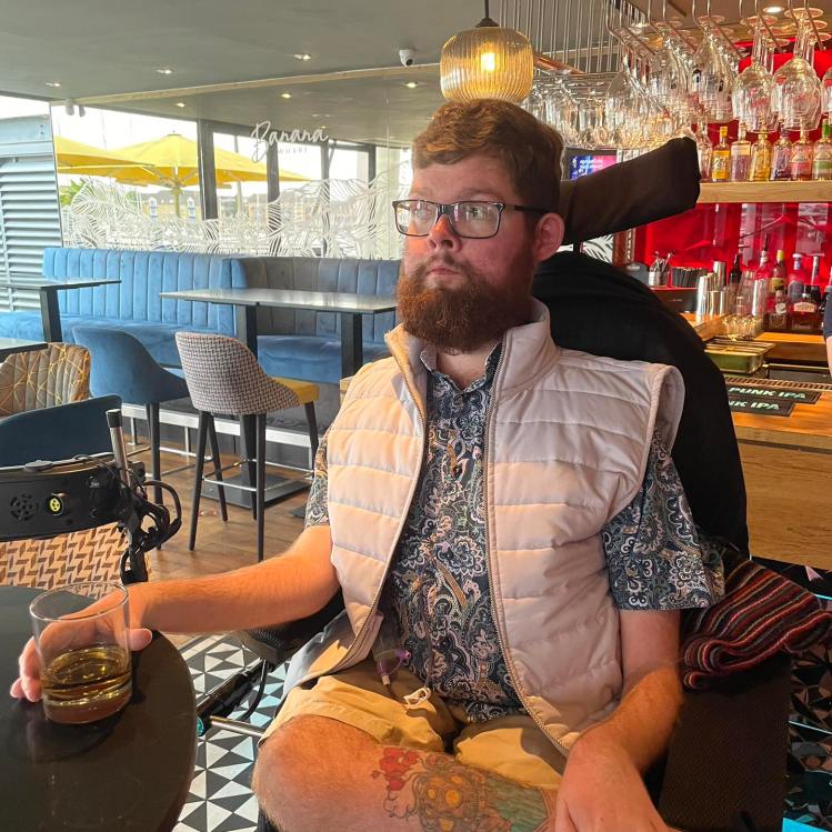 Photograph of Philip, who is wearing a white coloured Gilet and a patterned shirt. Phillip is using a wheelchair and is sat holding a drink in his right hand
