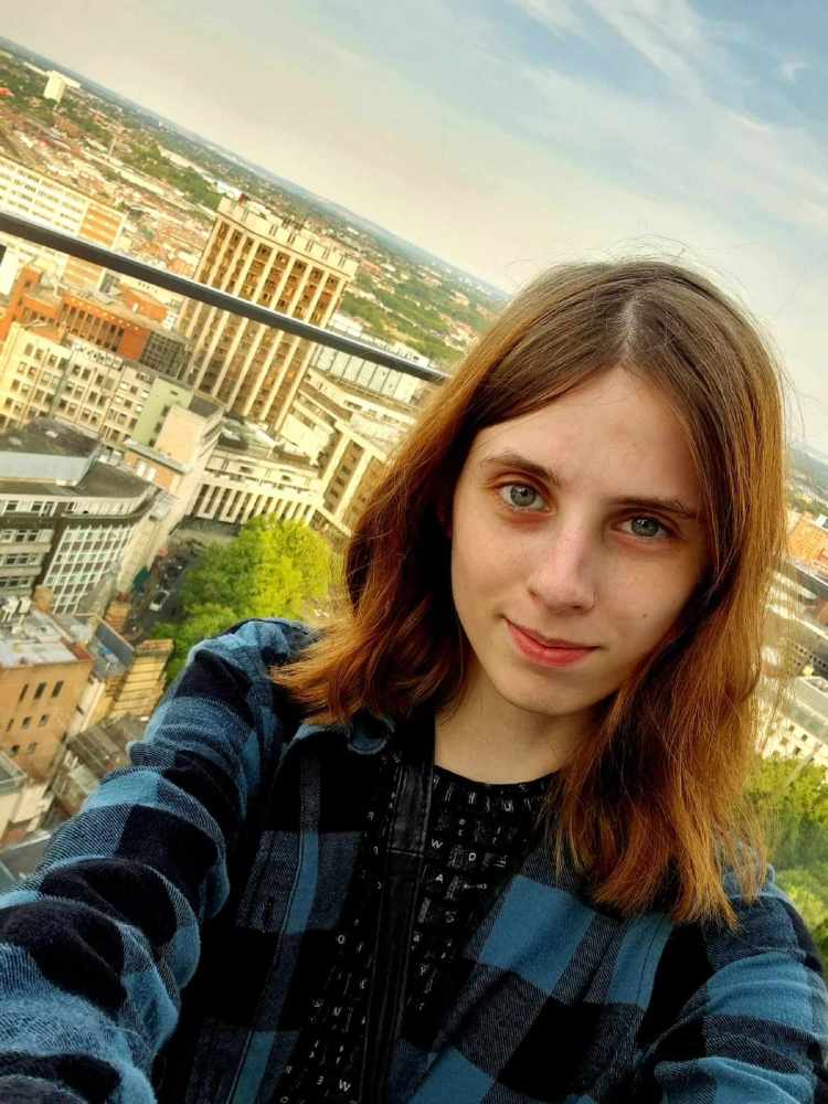 Photograph of Weronika, a white woman with shoulder length brown hair, who is wearing a checked shirt and is photographed outside, overlooking a city landscape
