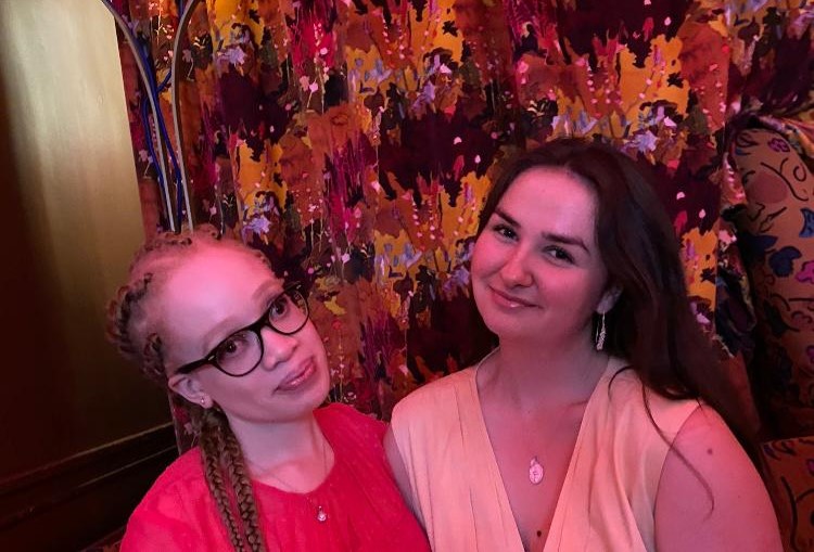 Rebecca, left, with black glasses and beautiful braided hair, smiling in a smart red dress. Kirstie, next to her on the right, with long pearl-design earrings and a silver necklace and a v-neck yellow dress. They are sat down, at the camera, with a light behind them and a wall with a dark leaf-like background.
