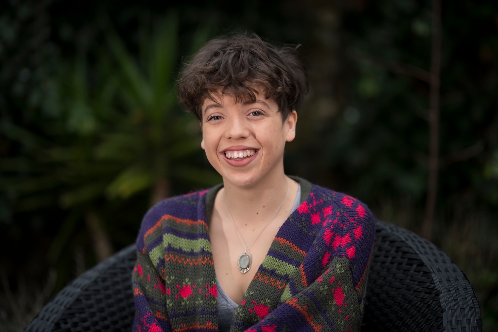 Photograph of Lucy, a white woman with short dark brown curly hair. Lucy is wearing a brightly coloured cardigan and is smiling towards the camera