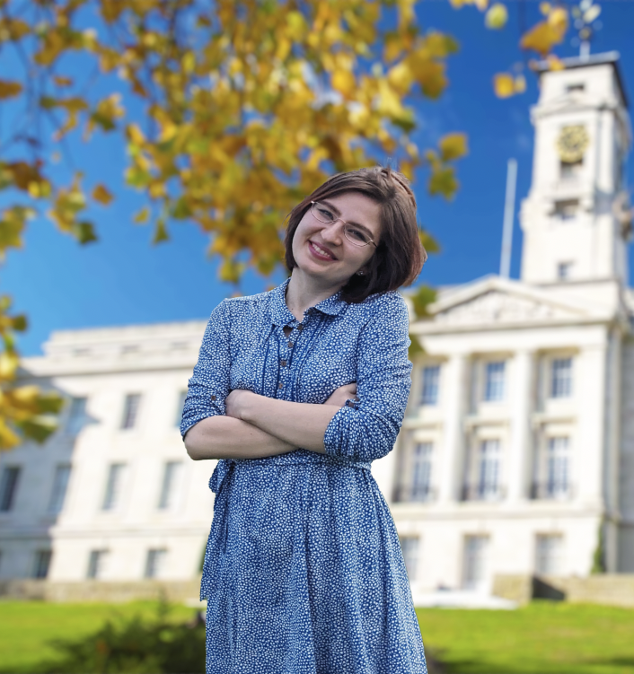 Maria is a white woman with short brown hair who is stood outside a white, grand building. Maria has her arms crossed and is holding her head to the side, looking at the camera. 