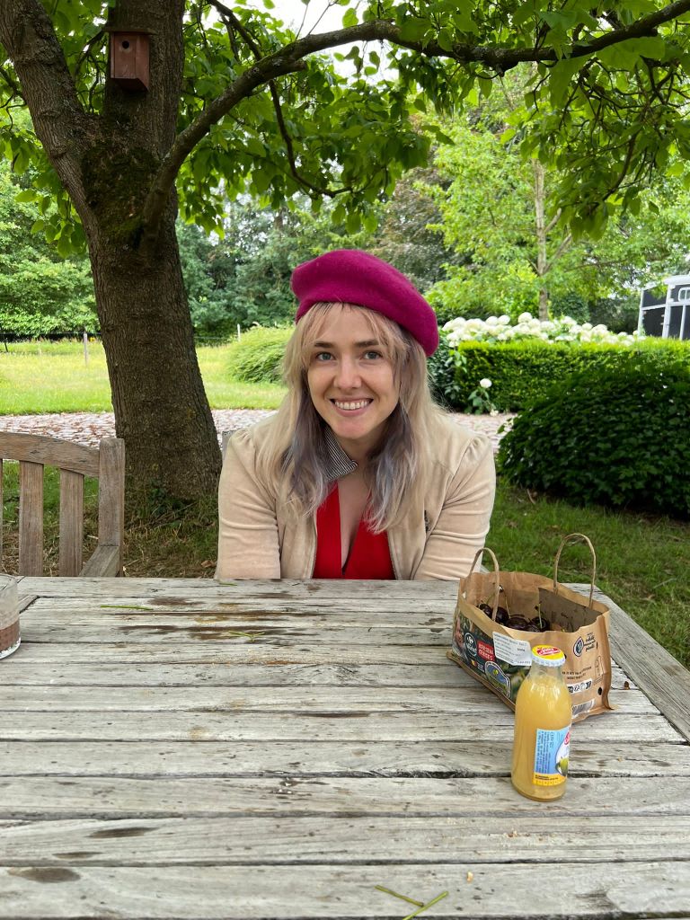 Photograph of Laura, a white woman with long blonde hair. Photographed outside, sat at a picnic table under a tree. Laura is wearing a red beret style hat and is smiling widely to the camera