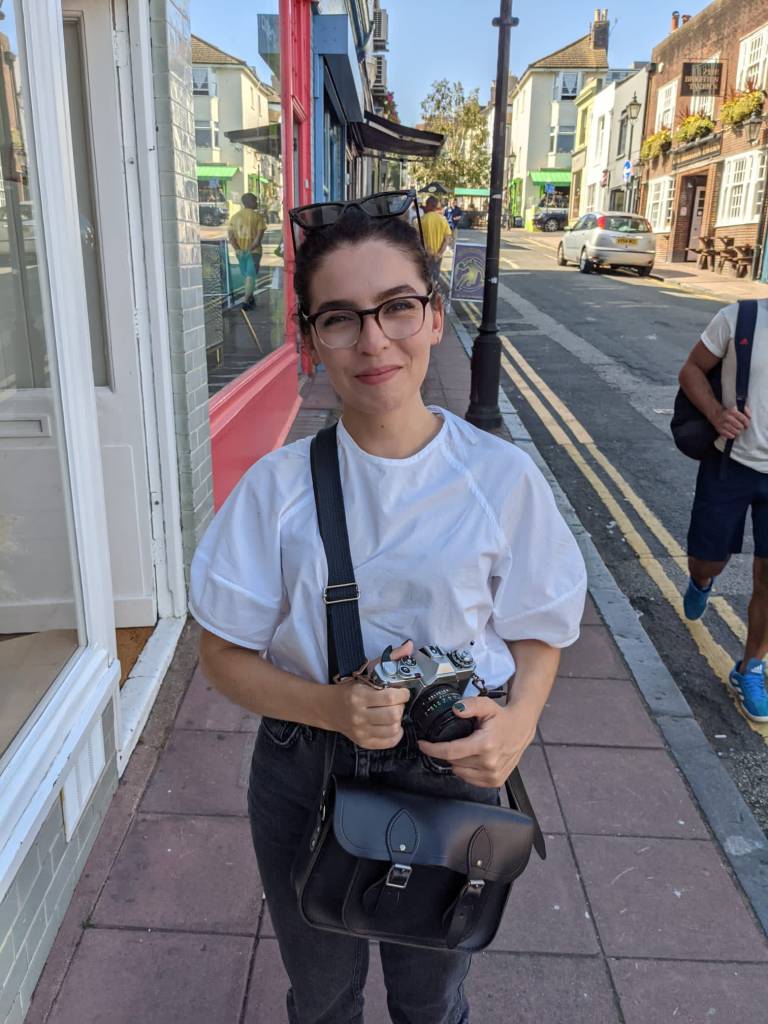 A colour photograph of Maera, a white woman with dark hair tied up away from her face. Maera is wearing a white t-shirt, is stood ourside on a street pavement and is holding a camera in her hand. 