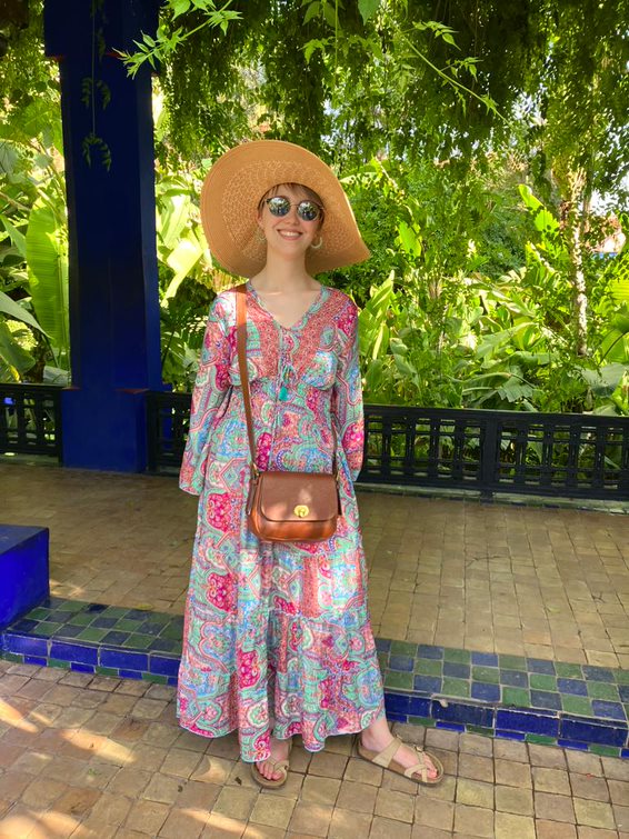 Photograph of Rhiannon, a woman wearing a long brightly printed dress, sunglasses and a summer hat. Rhiannon is smiling towards the camera, with her head tilted to one side. The weather is sunny. 