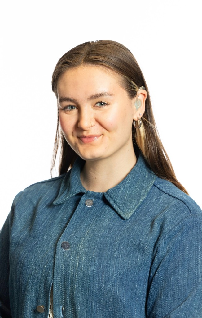 A professional photograph of Sian, a white woman with long brown hair who is smiling towards the camera. Sian has a nose piercing, hooped earrings and a hearing aid is visible in her left ear. 