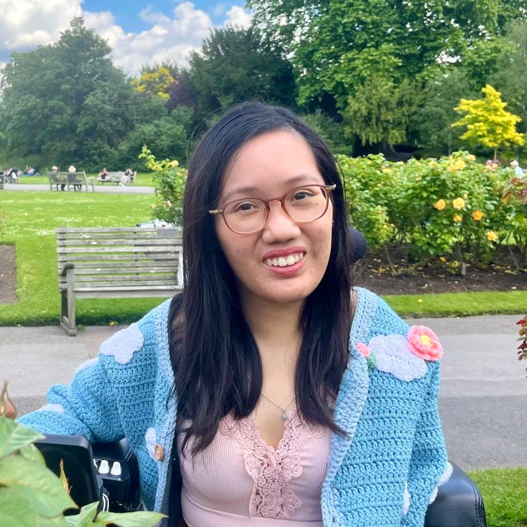 Photograph of Alicia Loh, an asian woman with long black hair, wearing glasses and smiling at the camera. Alicia is a wheelchair user and is photographed outside in a park. 